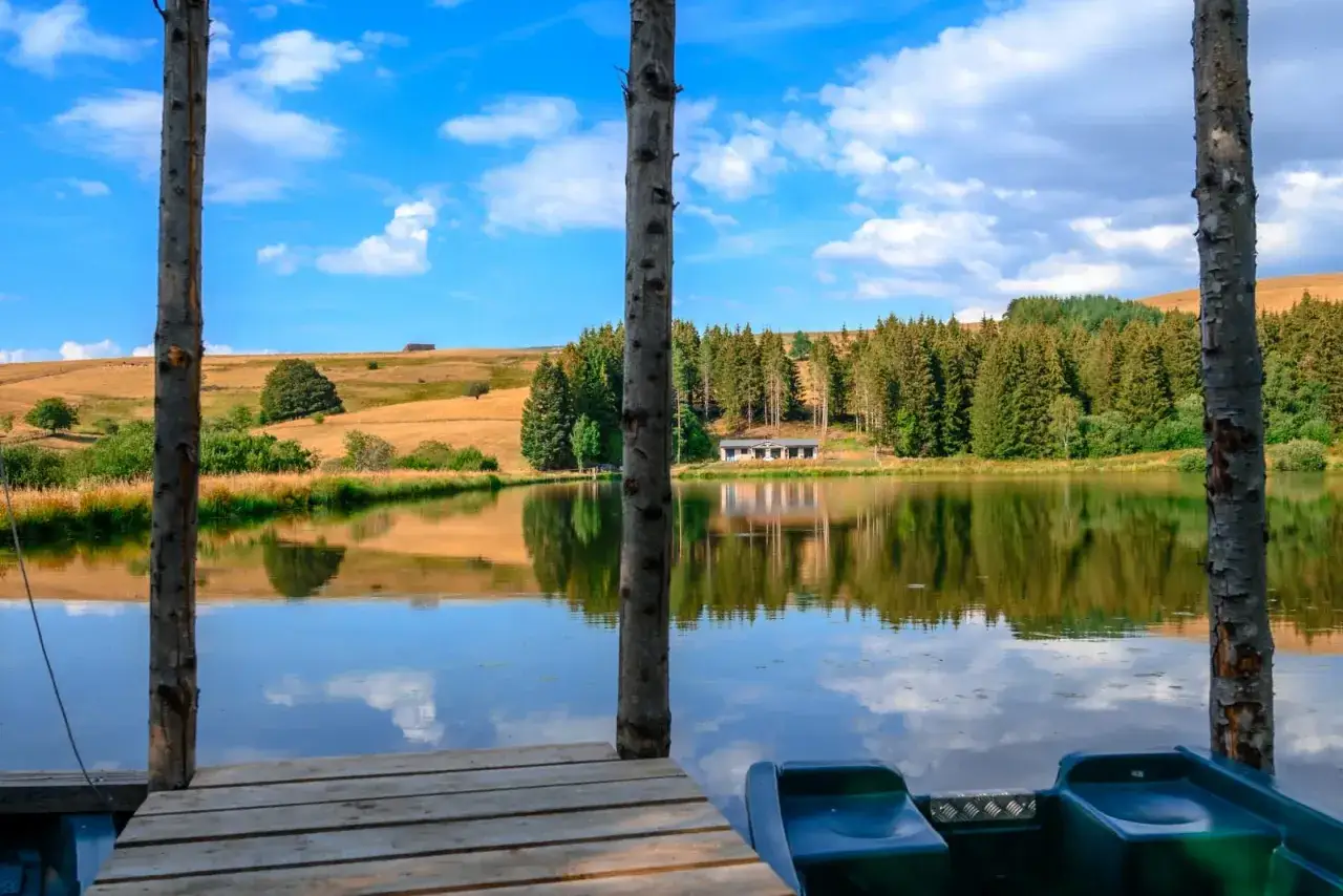 Vue sur un lac paisible, une maisonnette et une forêt dense, évoquant la petite Mongolie auvergnate sous un ciel bleu.