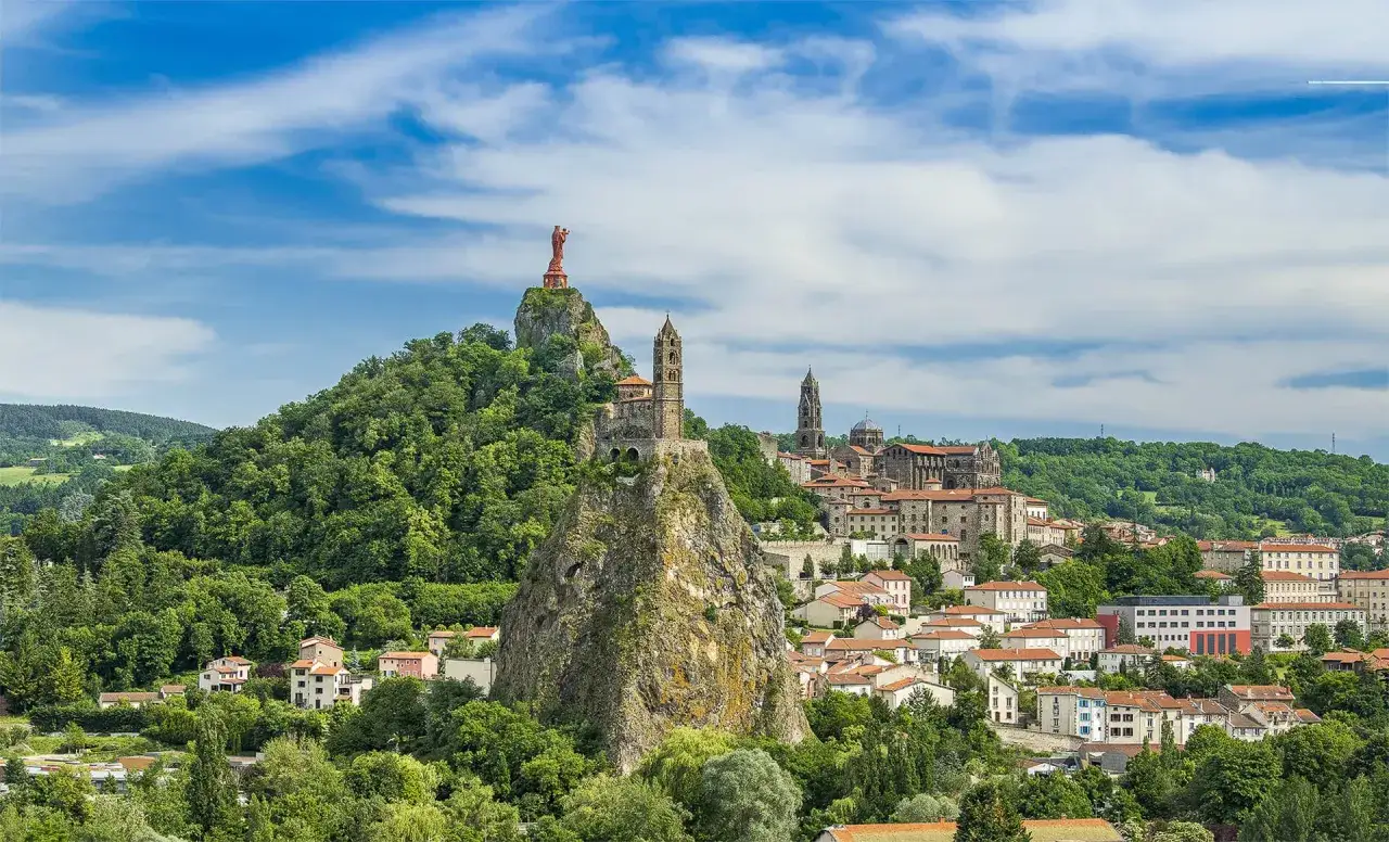 Vue panoramique de monuments emblématiques, dont la statue de Notre-Dame de France et la cathédrale, sur des pitons rocheux. Les incontournables autour du Puy-en-Velay sous un ciel bleu.