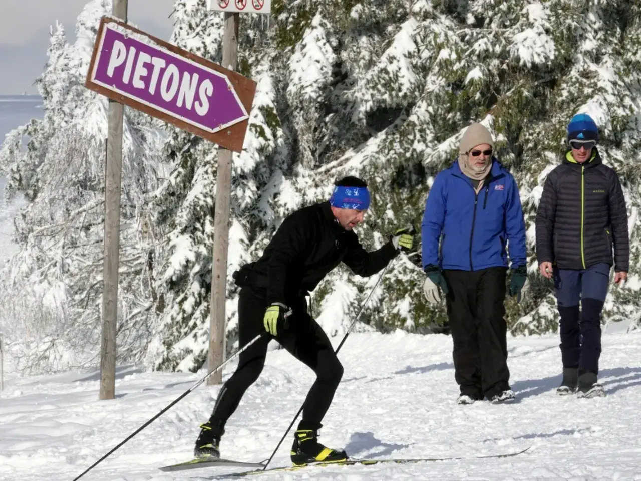 Trois hommes profitent du ski de fond sur le Champ du Feu. Un skieur avance, tandis que deux autres marchent. Un panneau indique