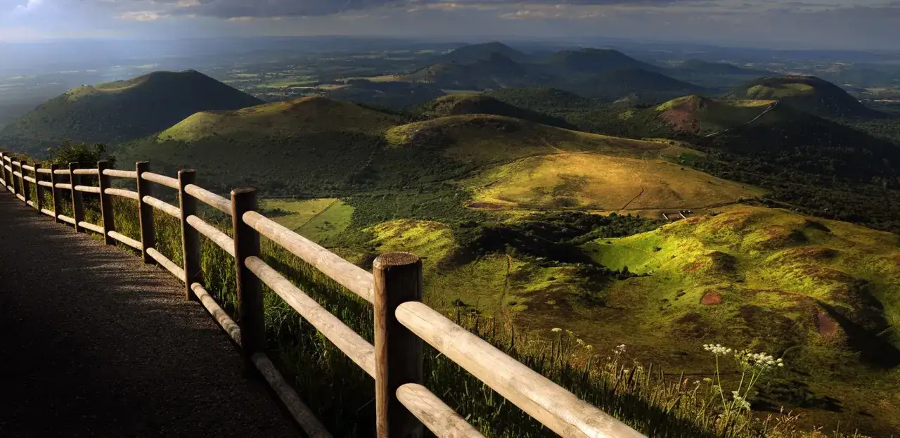 Vue panoramique sur les volcans du Puy Cantal, avec une clôture en bois au premier plan et des collines verdoyantes baignées de soleil.