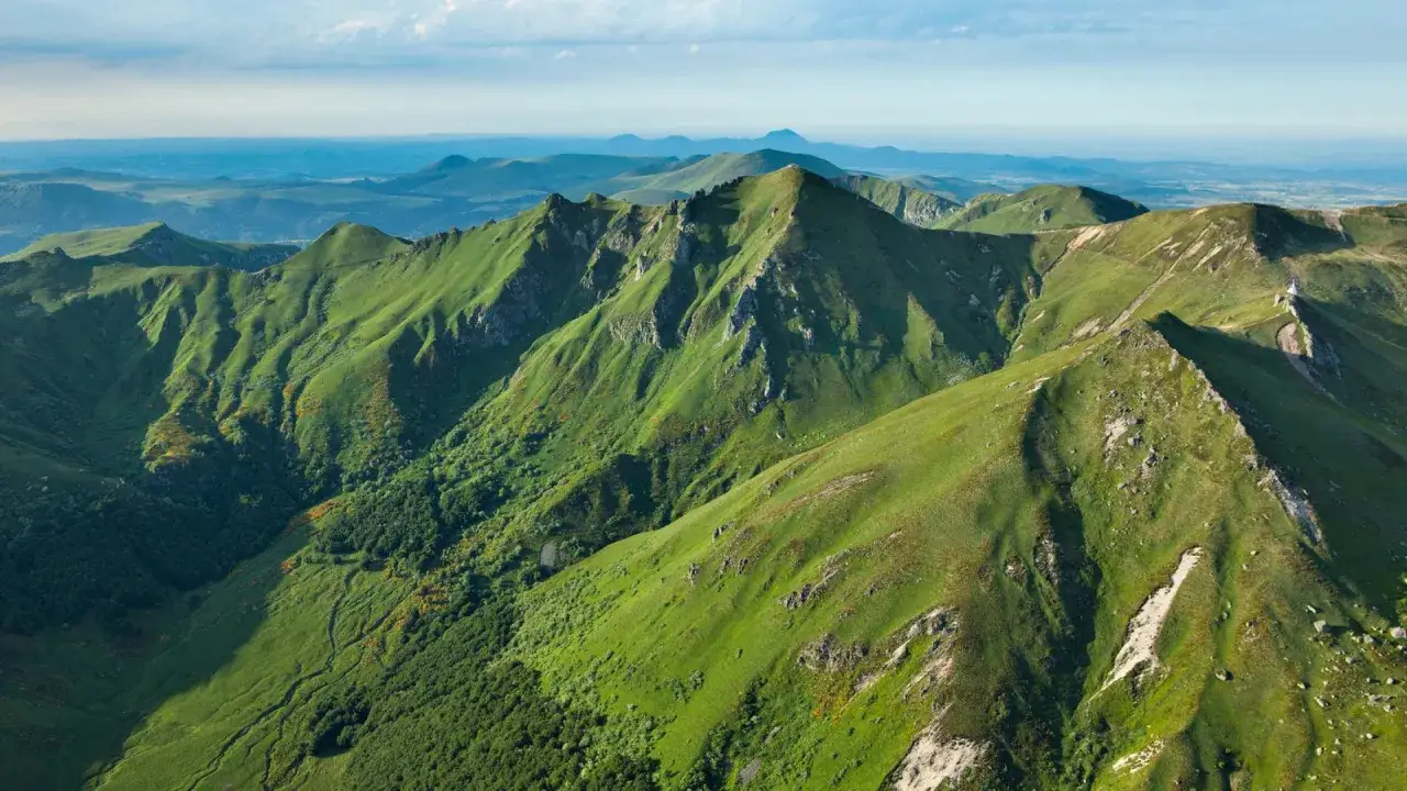 Vue a&eacute;rienne des montagnes verdoyantes du Massif Central, avec un petit b&acirc;timent blanc au sommet du plus haut sommet.