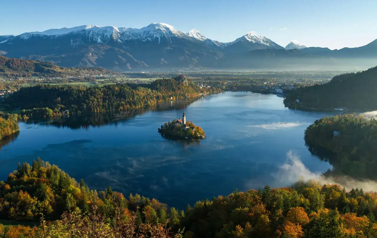 &Icirc;le avec &eacute;glise sur un lac bleu, entour&eacute;e de for&ecirc;ts automnales et de montagnes enneig&eacute;es. Un paysage incontournable en Slov&eacute;nie.