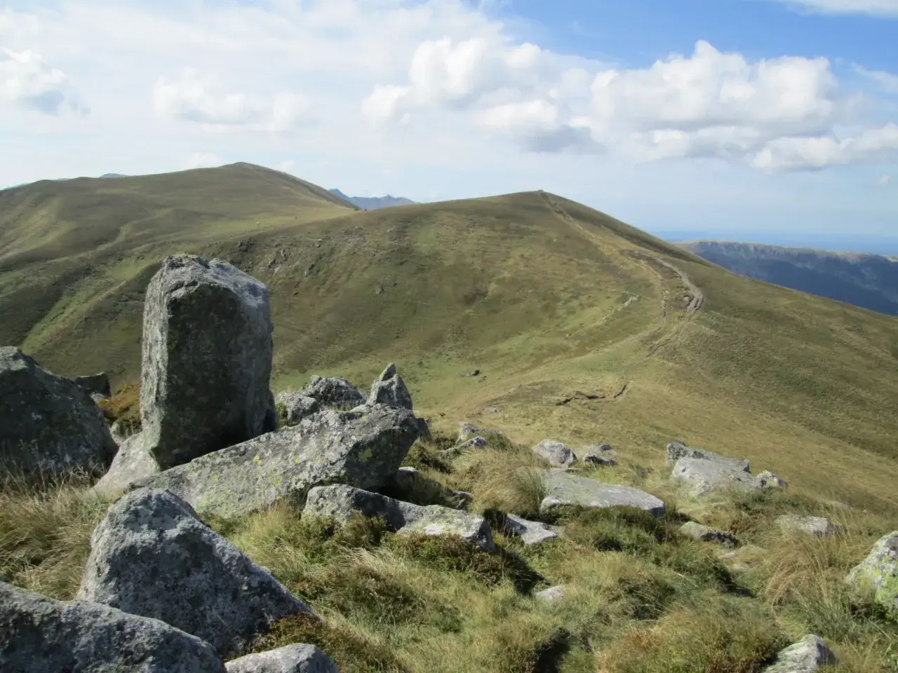 Un sentier serpente sur une colline verdoyante, menant vers un sommet où se dresse une croix morand. Des rochers parsèment le paysage.