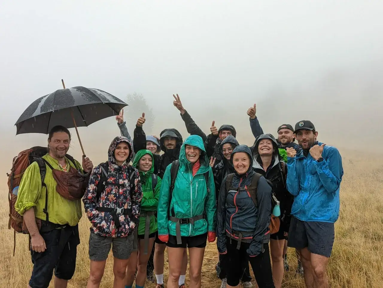 Groupe de randonneurs souriants sous la pluie, &eacute;quip&eacute;s de vestes imperm&eacute;ables et d'un parapluie, prouvant la norme IPX8 de leur mat&eacute;riel.