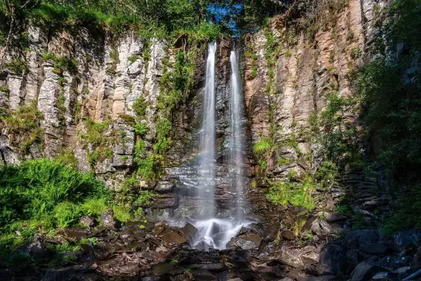 La plus belle cascade du Puy-de-Dôme, une chute d'eau double tombant sur des rochers escarpés, entourée de verdure luxuriante. Un arc-en-ciel apparaît dans la brume.