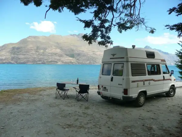 Camping-car blanc au bord d'un lac turquoise, table et chaises dressées. Un décor parfait pour un budget nouvelle zélande.