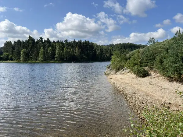 Magnifique paysage pour un tour du lac de Naussac à vélo. Eau calme, plage de galets et forêt dense sous un ciel bleu et nuageux.