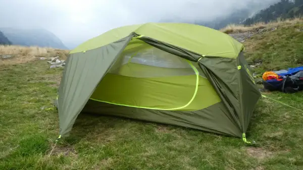 Tente verte et jaune, équipement de camping Nemo, installée sur une pelouse avec des montagnes brumeuses en arrière-plan.