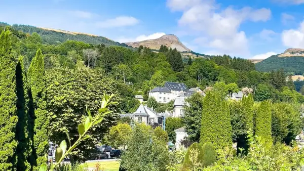 Vue d'un village niché dans une vallée verdoyante, avec des maisons aux toits gris et un grand bâtiment blanc. Au loin, une montagne imposante se dresse sous un ciel bleu. Un banne ordanche, peut-être une tente ou un auvent, est visible dans le paysage.