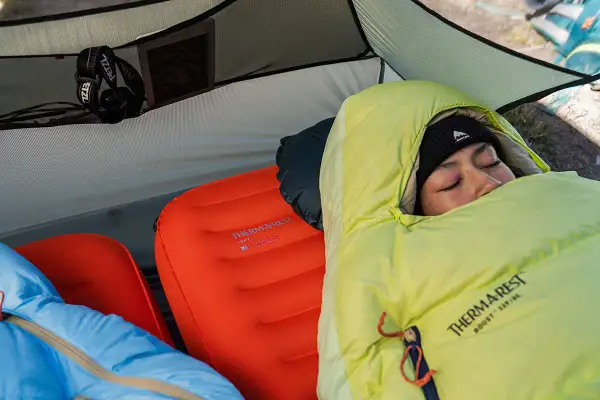 Une personne dort dans une tente sur deux matelas superposés. Le matelas du bas est orange, celui du dessus est bleu.