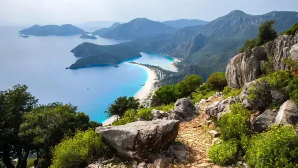 Sentier rocailleux du Lycian Way surplombant une baie turquoise et une plage de sable blanc bordée de montagnes verdoyantes.