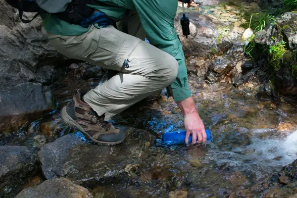 Randonneur remplissant une gourde dans un ruisseau, prêt à utiliser une pastille de chlore pour eau potable.