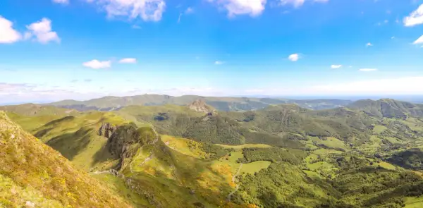 Magnifique panorama du Puy Mary, avec ses crêtes verdoyantes et ses vallées boisées sous un ciel bleu éclatant.