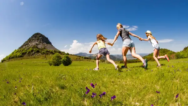 Une mère et ses deux filles courent main dans la main dans un champ fleuri, avec le Mont Gerbier de Jonc en toile de fond.