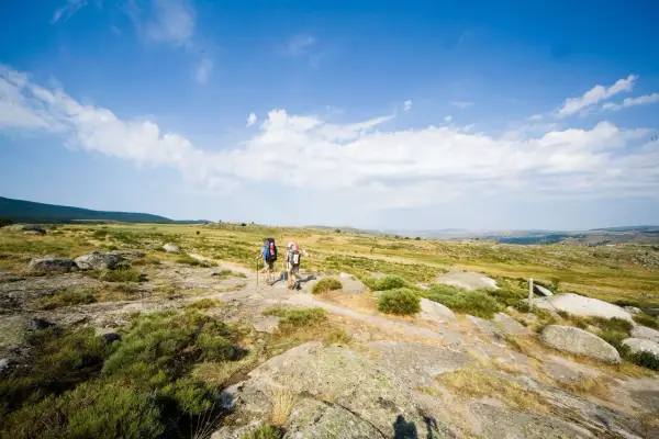 Deux randonneurs avancent sur un sentier rocailleux, sous un ciel bleu parsemé de nuages. Ils parcourent les gr 70 étapes, profitant du paysage.