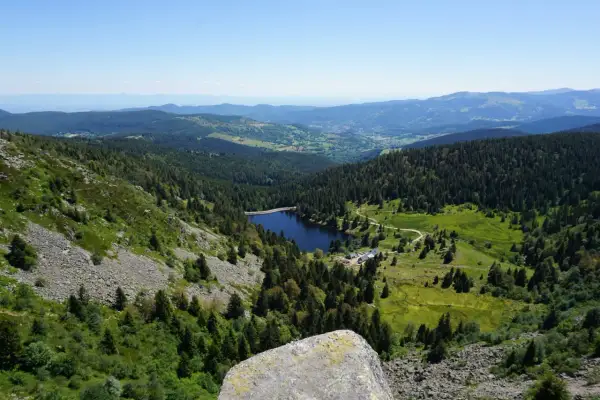 Vue panoramique sur un lac entouré de forêts denses, typique des 14 sommets des Vosges. Le ciel est clair et bleu.