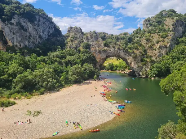 Les gorges du Chassezac, un pont naturel rocheux surplombe une rivière verdoyante où des kayakistes profitent du soleil sur une plage de galets.
