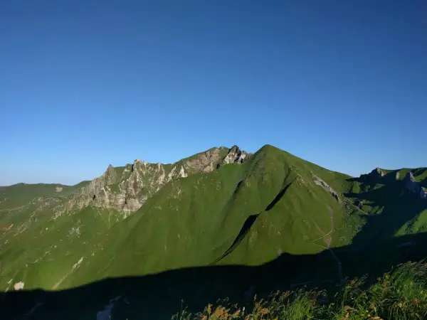Vue panoramique sur des montagnes verdoyantes sous un ciel bleu clair. Un sentier sinueux suggère une randonnée de **gr30 difficulté**.