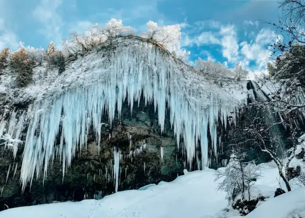 Impressionnante cascade gelée en Auvergne en hiver, avec des stalactites de glace bleutée tombant d'une falaise enneigée sous un ciel bleu et nuageux.