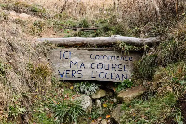 Panneau en bois aux **sources de la Loire** : "Ici commence ma course vers l'océan". Entouré d'herbes sèches et de rochers.