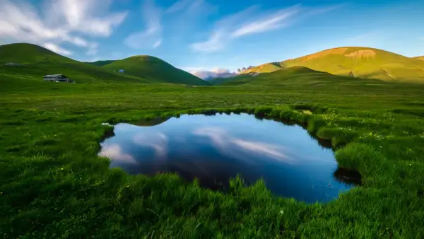 Un étang reflète le ciel et les collines verdoyantes, évoquant la petite Mongolie auvergnate. Un paysage paisible avec des nuages filant.