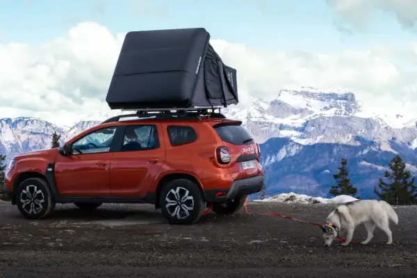 Un Dacia Duster orange avec tente de toit, prêt pour un bivouac Vanoise. Un husky renifle le sol, la neige et les montagnes en arrière-plan.