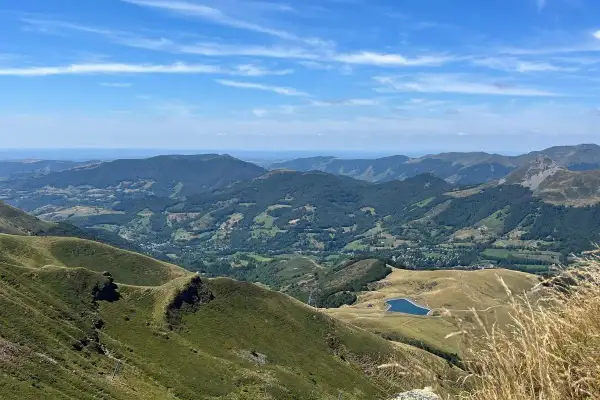 Vue panoramique des montagnes verdoyantes du Cantal, avec un lac bleu scintillant au loin. Le **Plomb du Cantal** domine l'horizon sous un ciel bleu parsemé de nuages.