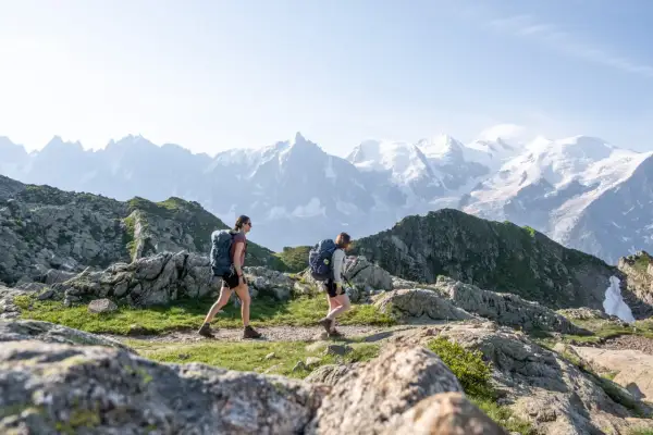 Deux randonneuses avec sacs à dos traversent un sentier rocheux, avec des montagnes enneigées en arrière-plan. Une randonnée accessible en train pour admirer ce paysage.
