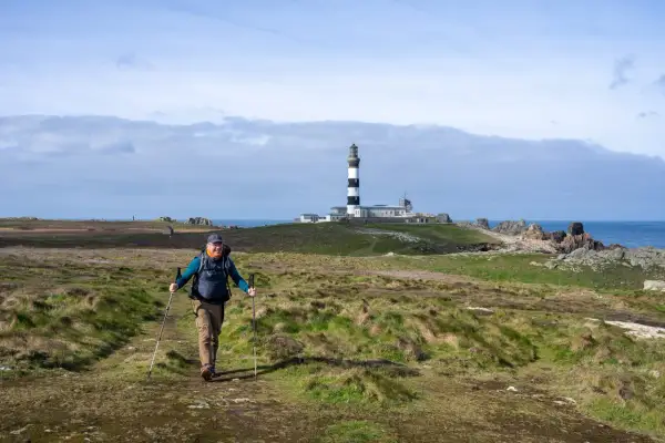 Un randonneur souriant marche sur un sentier côtier à Ouessant, avec le phare en arrière-plan.