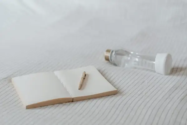 Un carnet ouvert avec un stylo doré et une gourde en verre. Idéal pour noter ses pensées et s'hydrater, sans enlever le goût plastique de la gourde.