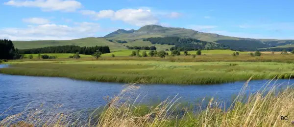 Vue paisible du lac Chauvet, entouré de prairies verdoyantes et de collines ondulantes sous un ciel bleu parsemé de nuages.