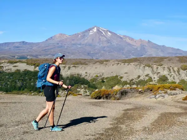 Randonneuse avec sac à dos et bâtons, portant une chaussure sans chaussette, marche sur un sentier rocailleux devant une montagne enneigée.