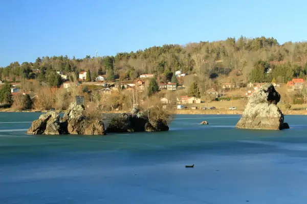Le lac de Cassière gelé, avec ses îlots rocheux et ses maisons sur la colline boisée, sous un ciel bleu clair.